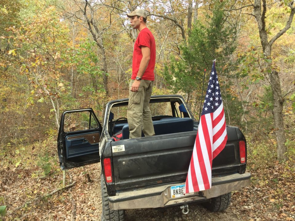 Man in red shirt standing in black pickup truck with American flag.