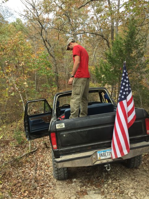 1979 Ford Bronco - A man in a red shirt and khaki pants stands in the bed of a black Ford Bronco with the driver's side door open. An American flag is attache…