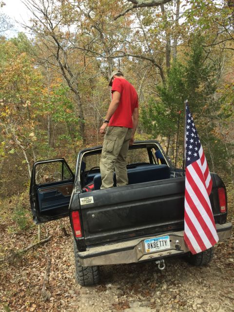 1979 Ford Bronco - A man in a red shirt and khaki pants stands in the bed of a black Ford pickup truck, which is parked on a dirt road with trees in the backg…
