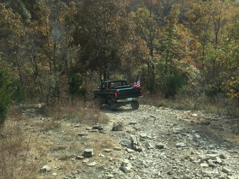 1979 Ford Bronco - A black 1970s Ford pickup truck with an American flag in the bed drives on a rocky, wooded trail during autumn.
