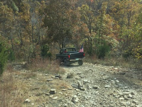 1979 Ford Bronco - A black 1979 Ford Bronco is parked on a rocky, wooded trail with an American flag in the bed.