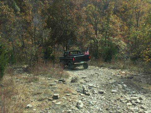 1979 Ford Bronco - A black 1979 Ford Bronco is parked on a rocky, wooded trail during the fall. An American flag is attached to the rear of the vehicle.