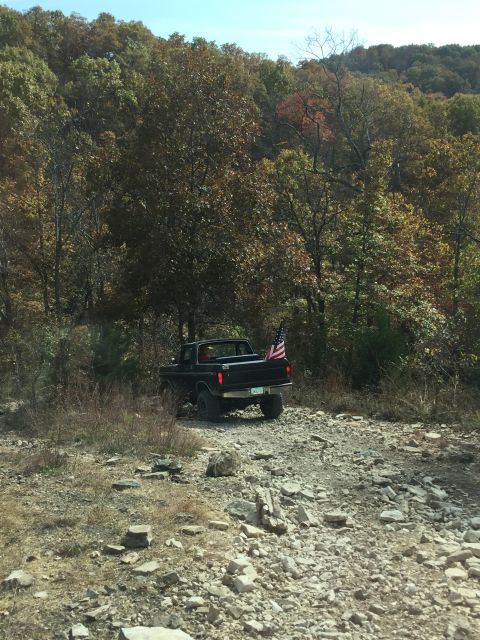 1979 Ford Bronco - A black pickup truck with an American flag attached to the tailgate is parked on a rocky, unpaved road with trees in the background.