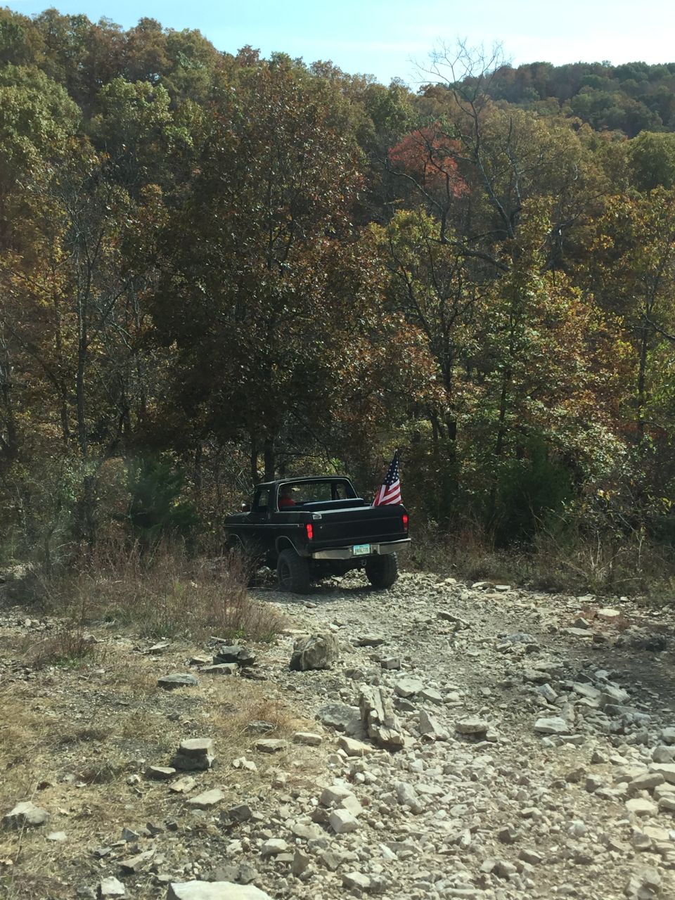 Black pickup truck with American flag on a rocky road in a wooded area.