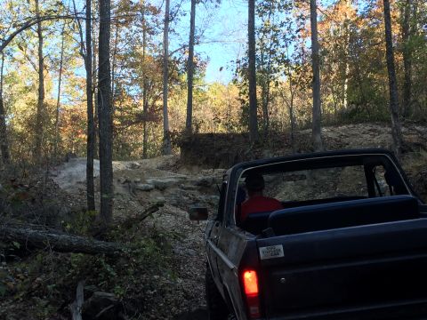 1979 Ford Bronco - A dark-colored, convertible Ford Bronco drives on a rocky, off-road trail through a forest with autumn foliage. A sticker on the rear bumpe…