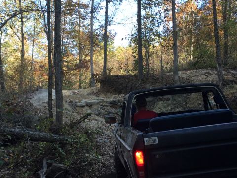 1979 Ford Bronco - The rear view of a dark-colored, convertible-top Ford Bronco driving on a rocky, wooded trail during autumn. A sticker on the tailgate read…