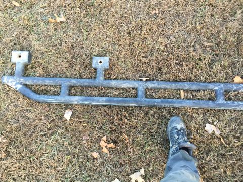 1979 Ford Bronco - A black metal rock slider with two mounting brackets is laid out on dry grass. A person wearing camouflage pants and a hiking boot stands n…