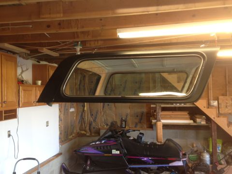 1979 Ford Bronco - A black camper shell with a large window is suspended from the ceiling of a garage. Below it sits a snowmobile with purple and black graphi…