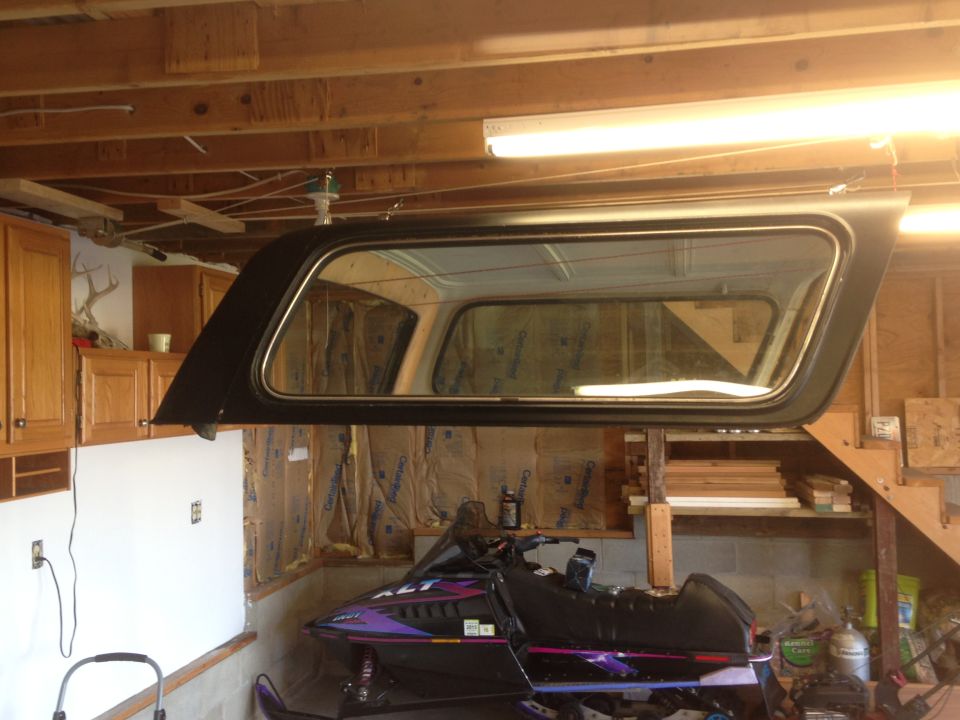 Black camper shell with window suspended above a snowmobile in a garage.