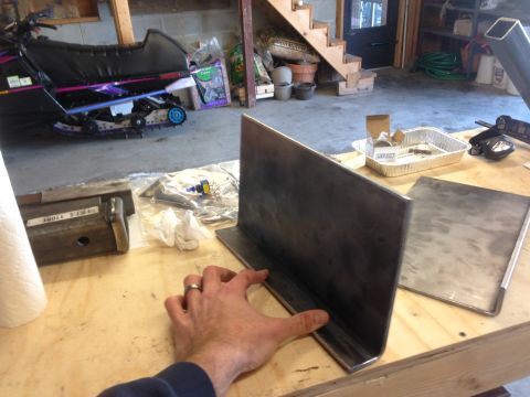 1979 Ford Bronco - A hand is holding a piece of metal that is L-shaped, with another flat piece of metal to its right on a wooden workbench. In the background…