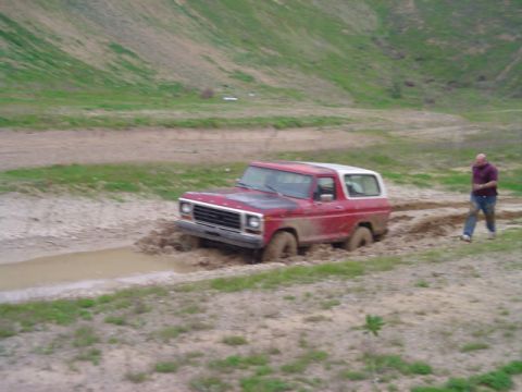 1978 Ford Bronco - A red and white 1978 Ford Bronco navigates a muddy off-road trail, with a person observing from the side.