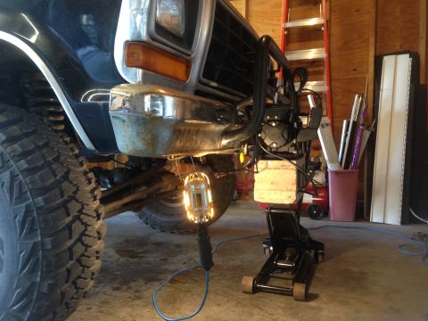 1979 Ford Bronco - A close-up, low-angle view of the front bumper and tire of a dark-colored vehicle, possibly a Ford Bronco, being worked on in a garage. A f…