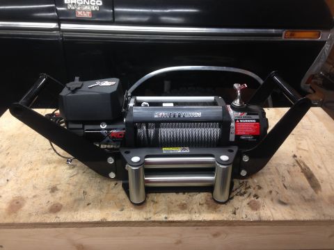 1979 Ford Bronco - A black Smittybilt XRC winch with a steel cable is mounted on a black metal frame. The winch is resting on a wooden surface in front of the…