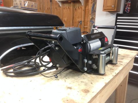 1979 Ford Bronco - A black winch with a steel cable is mounted on a wooden workbench in front of a black vehicle. The winch has red and silver accents and is…