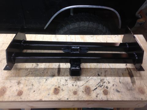 1979 Ford Bronco - A black, custom-fabricated trailer hitch receiver is shown mounted on a wooden workbench, with the rear fender and tire of a black vehicle…