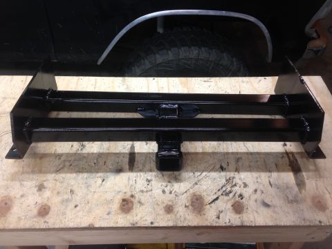 1979 Ford Bronco - A black, custom-fabricated trailer hitch receiver is mounted on a wooden workbench, with the rear fender and tire of a black vehicle visibl…