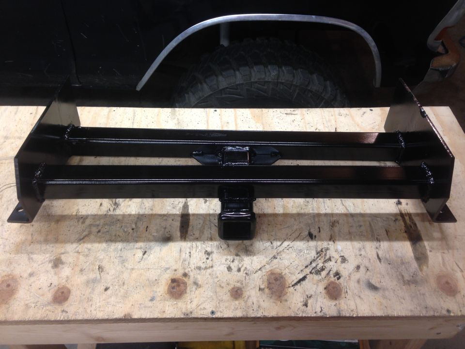 Black trailer hitch receiver mounted on a wooden surface, with a black vehicle's tire and fender in the background.