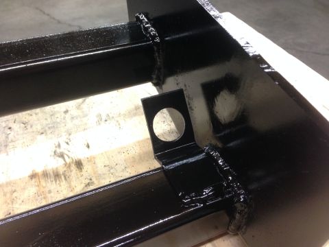 1979 Ford Bronco - A close-up view of a black metal part, likely a trailer hitch receiver, with visible welds and a circular hole. The part rests on a wooden…