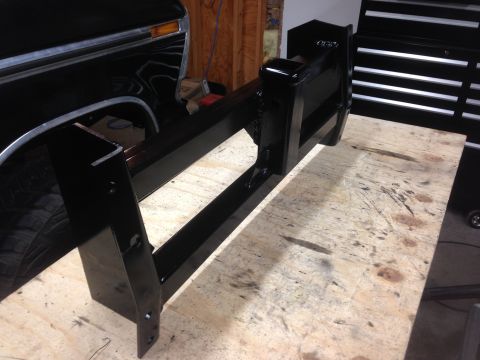 1979 Ford Bronco - A black metal fabrication, possibly a hitch or trailer attachment, rests on a wooden workbench in front of a black Ford Bronco. The part is…
