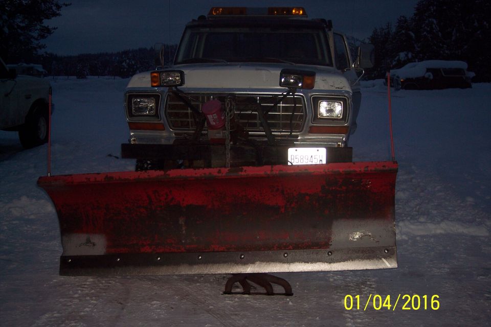 Front view of a vintage Ford pickup truck with a snow plow attached, in a snowy environment.