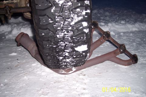 1973 Ford F-250 - A close-up view of a tire with snow on it, resting on a rusty exhaust pipe and part of a snow chain. The scene is set in a snowy environmen…