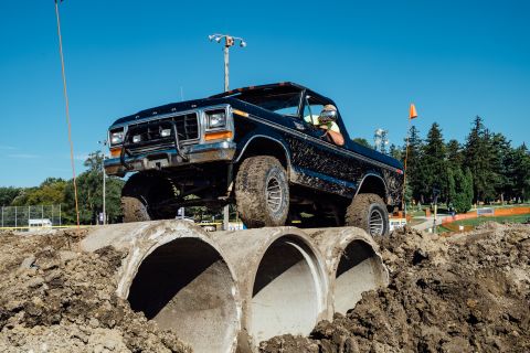 1979 Ford Bronco - A black Ford pickup truck with large off-road tires is driving over a series of large concrete pipes on a dirt track. The truck is covered…