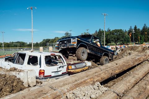 1979 Ford Bronco - A black 1979 Ford Bronco is driving over a white SUV and a brown car, navigating a course made of logs and dirt. The Bronco is lifted and h…