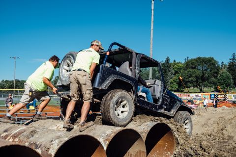 1979 Ford Bronco - Two men in bright yellow shirts push a black Jeep Wrangler as it navigates over large metal pipes on a dirt obstacle course.