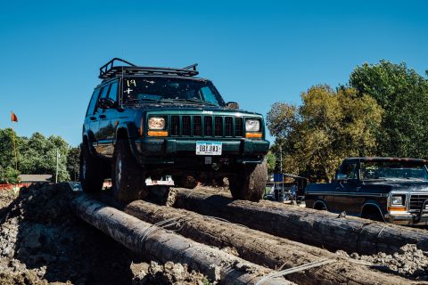 1979 Ford Bronco - A dark green Jeep Cherokee XJ is navigating an obstacle course made of large logs, with a black Ford pickup truck visible behind it.