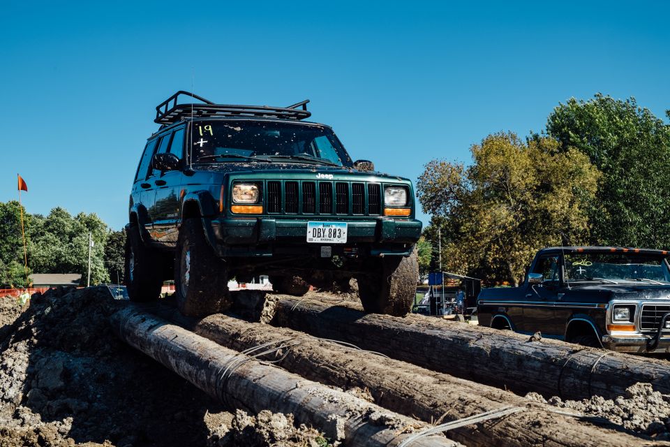 Green Jeep Cherokee on a log obstacle course, with a black Ford pickup behind it.