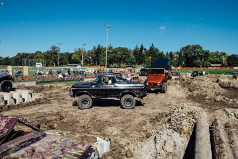 1979 Ford Bronco - A black 1970s Ford Bronco is shown in a muddy off-road course, with an orange Jeep Wrangler behind it. Spectators are visible in bleachers…