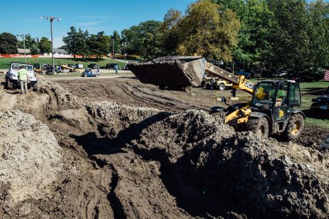 1979 Ford Bronco - A yellow JCB telehandler is moving a large amount of dirt in a muddy construction site. In the background, a white Jeep Wrangler is parked.