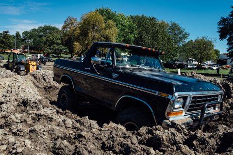 1979 Ford Bronco - A black 1979 Ford Bronco is stuck in a deep mud pit, with a yellow tractor visible in the background. The Bronco has large tires and a modi…