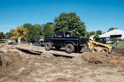 1979 Ford Bronco - A black Ford Bronco is driving over a log obstacle on a dirt course, with a yellow skid steer loader in the background. The Bronco has larg…