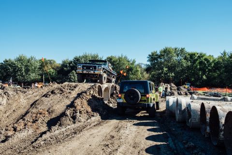 1979 Ford Bronco - A black 1979 Ford Bronco is driving up a dirt mound on an off-road course, with a yellow Jeep Wrangler following behind it. Several large c…