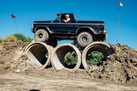 1979 Ford Bronco - A black 1979 Ford Bronco is driving over large concrete pipes on a dirt mound under a clear blue sky. Orange flags mark the obstacle course.