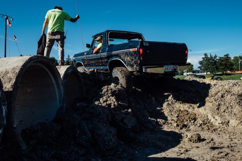 1979 Ford Bronco - A black 1970s Ford pickup truck with mud splattered on its sides navigates an obstacle course made of dirt mounds and large concrete pipes.…