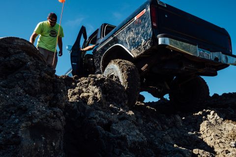 1979 Ford Bronco - A black, mud-covered pickup truck is navigating a steep, rocky, and muddy incline. A man in a lime green shirt stands next to the truck, ho…