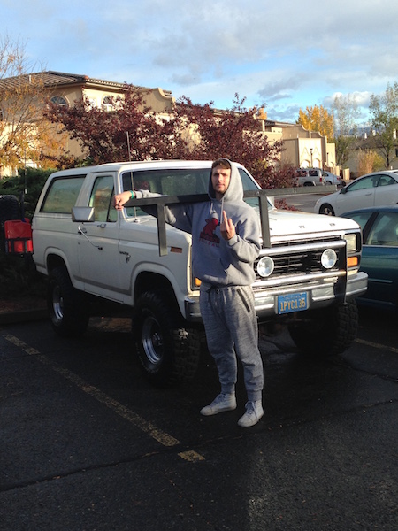 1981 Ford Bronco - A man in a grey hoodie and sweatpants stands in front of a lifted white 1980s Ford Bronco, holding a metal frame. The Bronco has large off-…