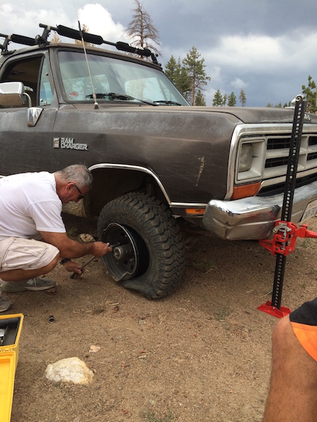 1981 Ford Bronco - A man is changing a flat tire on a brown Dodge Ramcharger. A red Hi-Lift jack is supporting the vehicle.