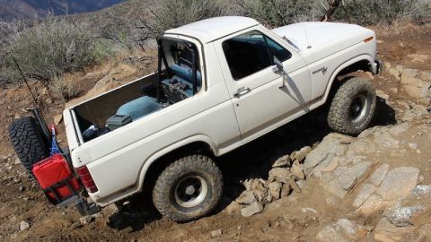 1981 Ford Bronco - A white 1981 Ford Bronco is shown from a high angle, navigating a rocky, off-road trail. The vehicle has large off-road tires, a spare tire…