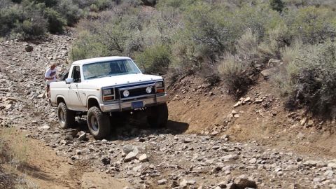 1981 Ford Bronco - A lifted white Ford pickup truck navigates a rocky, off-road trail with sparse vegetation on the hillsides. A person stands near the truck…