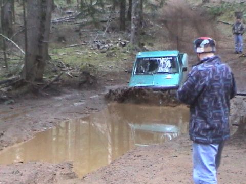 1994 Ford Explorer - A teal SUV navigates a deep mud puddle on a wooded trail, kicking up a large spray.