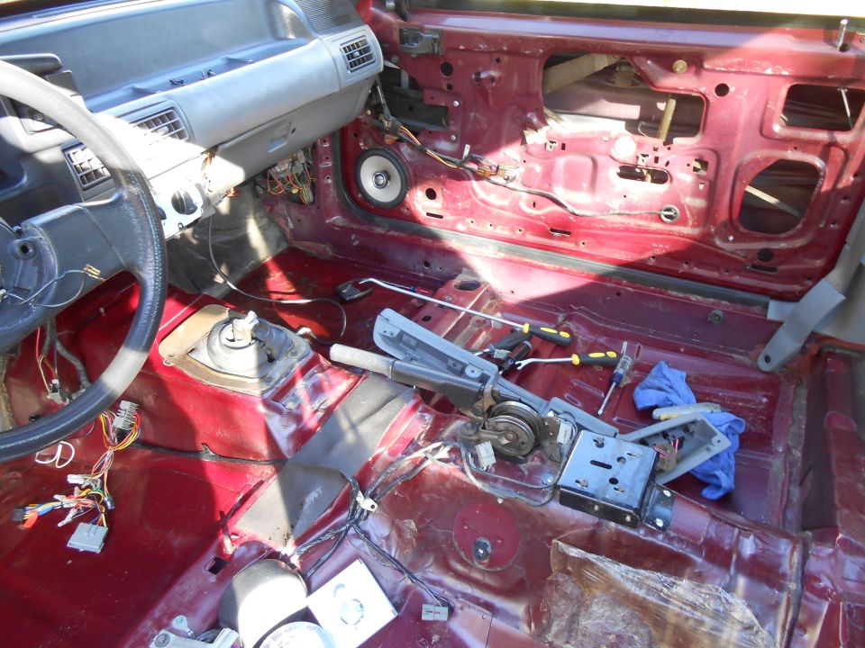 Disassembled car interior showing bare red metal, steering wheel, dashboard, and wiring.