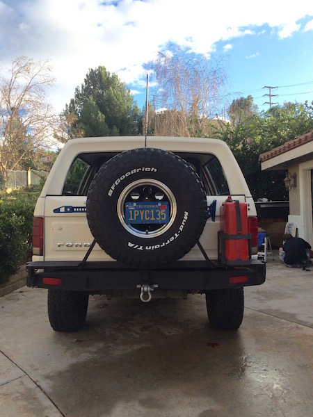 1981 Ford Bronco - The rear view of a white Ford Bronco is shown parked in a driveway. It has a spare tire mounted on the back, a black aftermarket bumper, an…