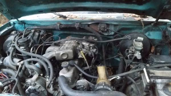 Engine bay of a teal 1982 Ford F-100 with hood open, showing hoses and engine parts.