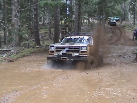 1994 Ford Explorer - A tan pickup truck navigates a muddy trail, kicking up a large spray of mud. Other vehicles and people are visible in the background.