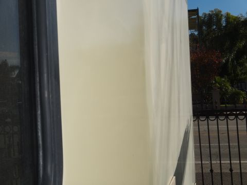 1995 Chevrolet P30 1 Ton - Step Van - A close-up side view of a cream-colored step van shows a dark window frame on the left and a black metal fence in front of a street and tre…
