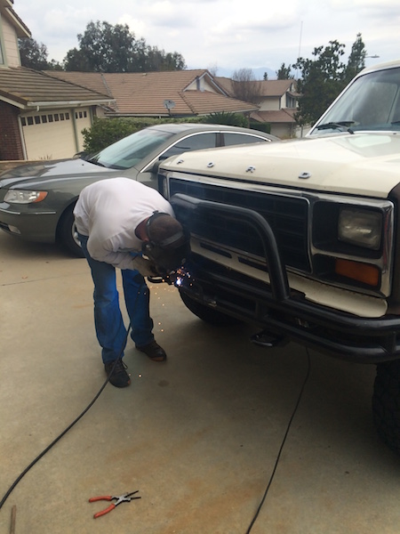 1981 Ford Bronco - A person wearing a white shirt and blue jeans is welding the front bumper of a white Ford truck. Sparks are flying from the welding point,…