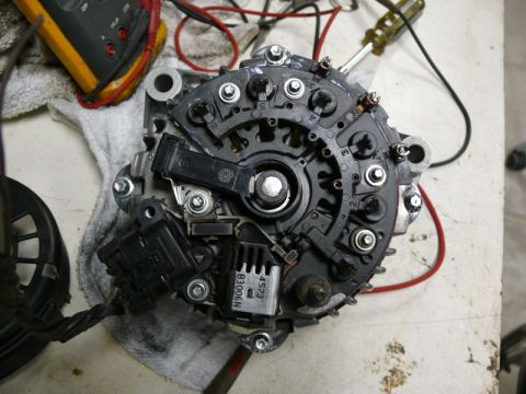 Overhead view of a disassembled car alternator with tools and multimeter in background.